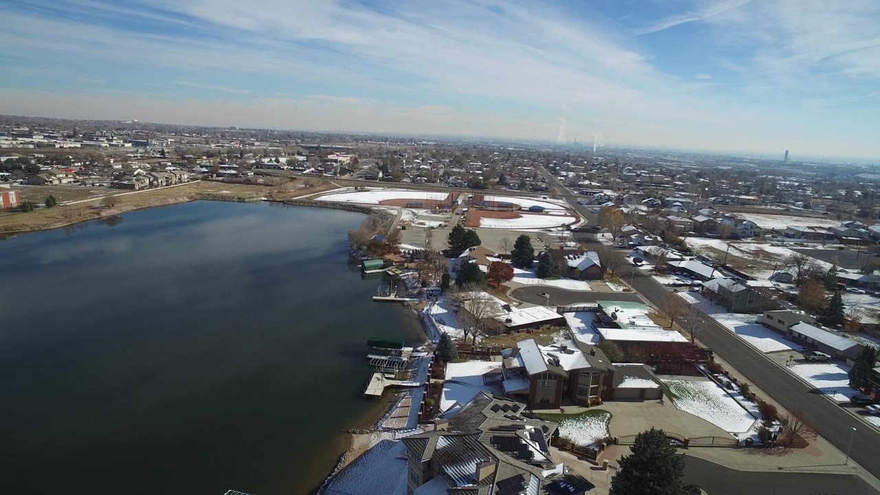 Hidden Lake, Colorado appears from above with smooth water across the frame, waterfront houses and docks near the lower edge, and suburban blocks stretching to the horizon, with no visible people.