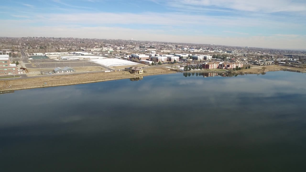 At Hidden Lake, Colorado, dark water fills the foreground while low industrial buildings and distant suburban blocks sit under a pale blue sky, with no visible people.