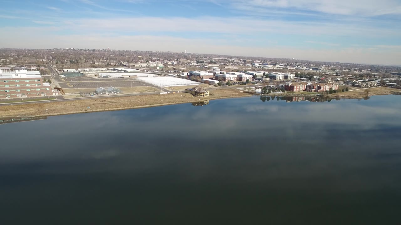 Wide drone view of lake water, shoreline buildings, and a distant residential area.
