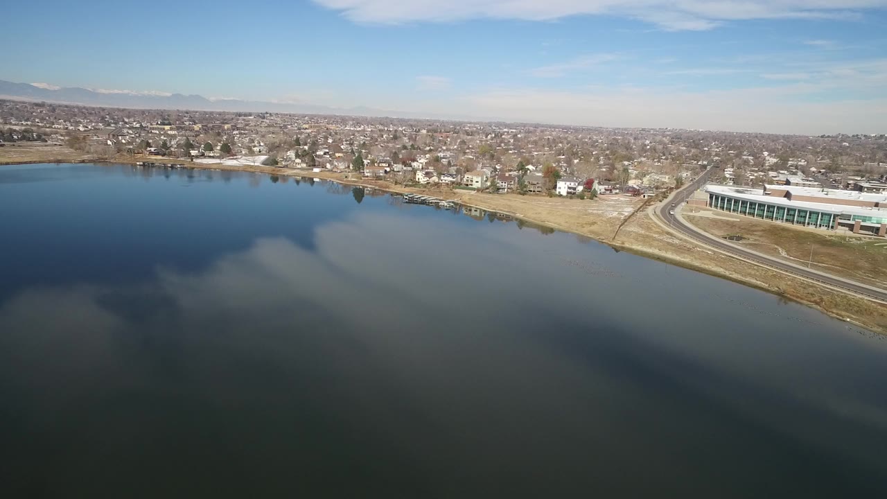 A high aerial frame over Hidden Lake, Colorado shows dark lake water with cloud reflections, shoreline homes, a road, a large building on the right, distant town blocks, mountains, and no visible people.