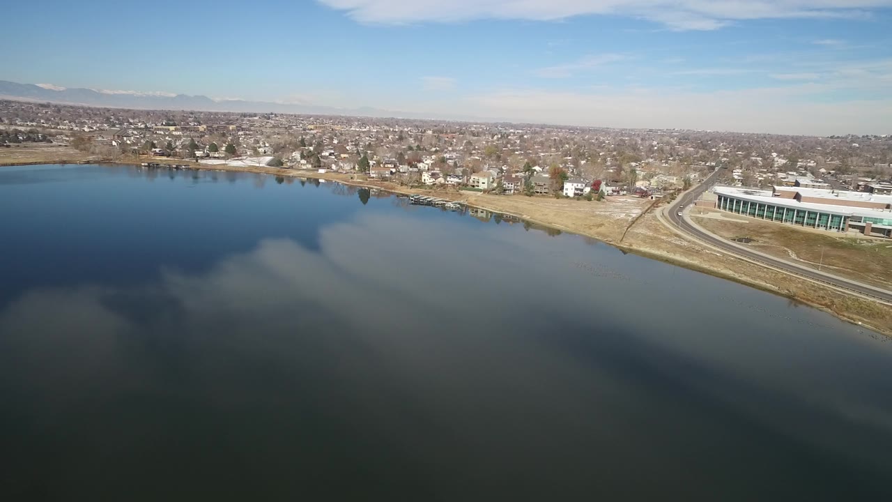 At 0:01, the Hidden Lake aerial holds a wide view of calm water, pale shoreline, small homes, a curving road, a large structure near the right edge, distant mountains, and no visible people.