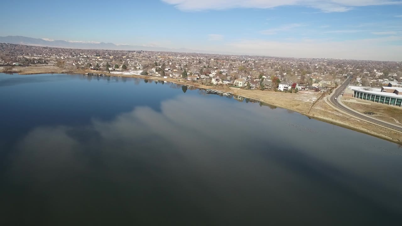 Wide aerial over a lake with houses along the shore and mountains in the distance.
