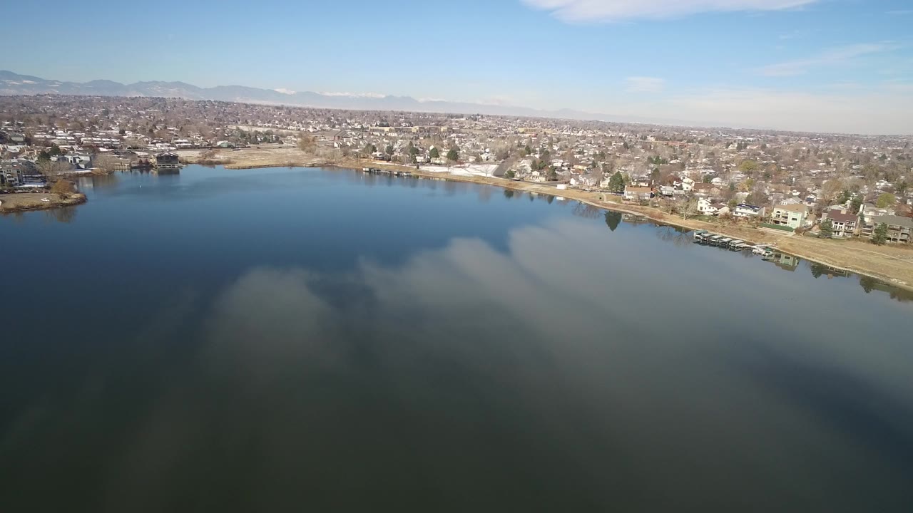 Aerial frame over Hidden Lake, Colorado with dark lake water in front, shoreline houses and leafless trees across the far bank, distant mountains on the horizon, and no visible people.