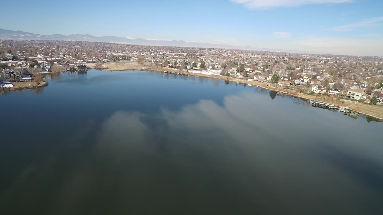 Lake shoreline with residential buildings, trees, and mountains under a clear sky.