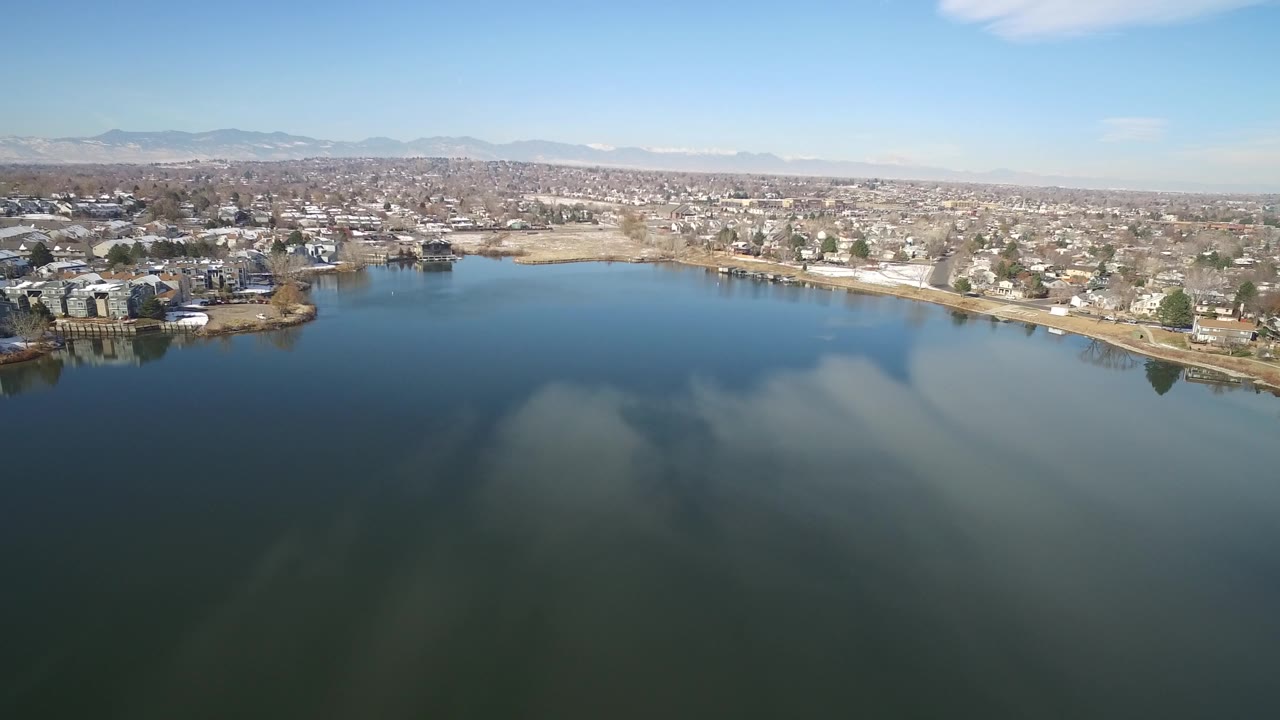 Aerial frame from Hidden Lake, Colorado, showing dark blue water with reflected clouds, clusters of homes along the shore, light snow on the ground, and no visible people.