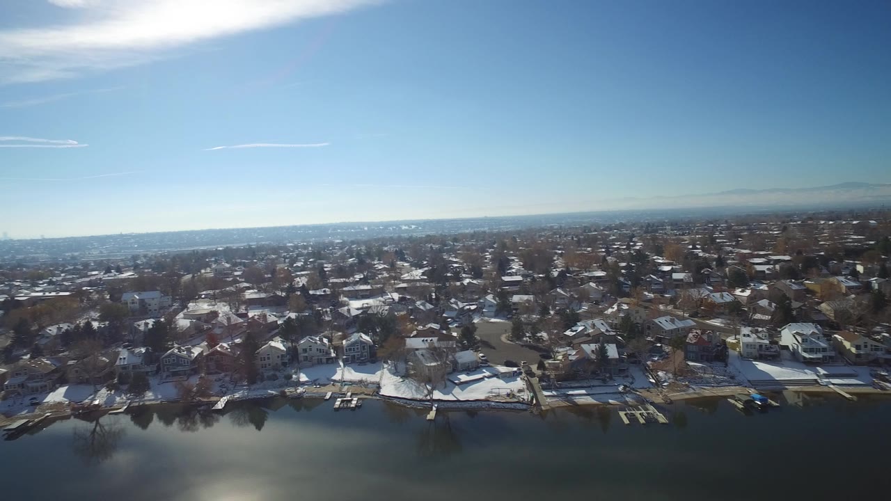 Elevated view across a lake in Hidden Lake, Colorado, showing shoreline homes, docks, snow patches, bare trees, a distant horizon, and no visible people.