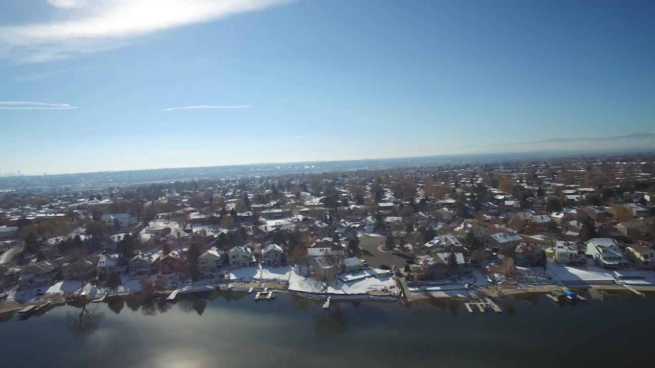 A wider elevated frame in Hidden Lake, Colorado, shows calm lake water below, a row of homes and piers along the shore, hazy distance, and no visible people.