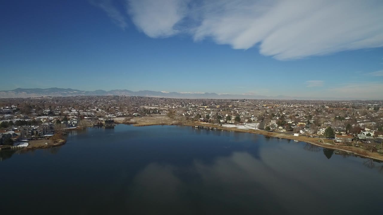 High aerial frame over Hidden Lake, Colorado, with dark lake water in the foreground, homes along the opposite shore, distant mountains, wispy cloud cover, and no visible people.