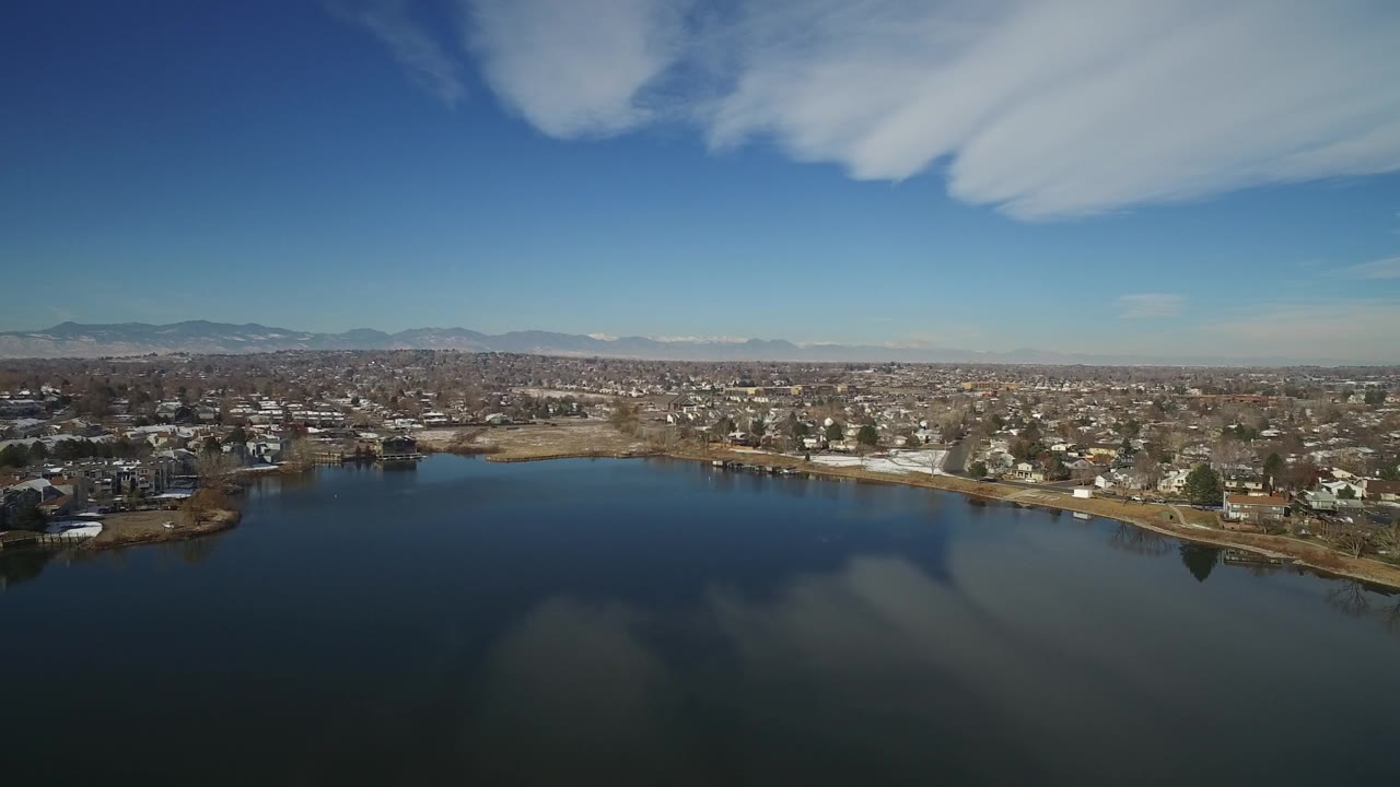 Aerial view in Hidden Lake, Colorado, showing broad lake water, a developed shoreline, clusters of houses, a flat suburban grid, distant mountains, and no visible people.