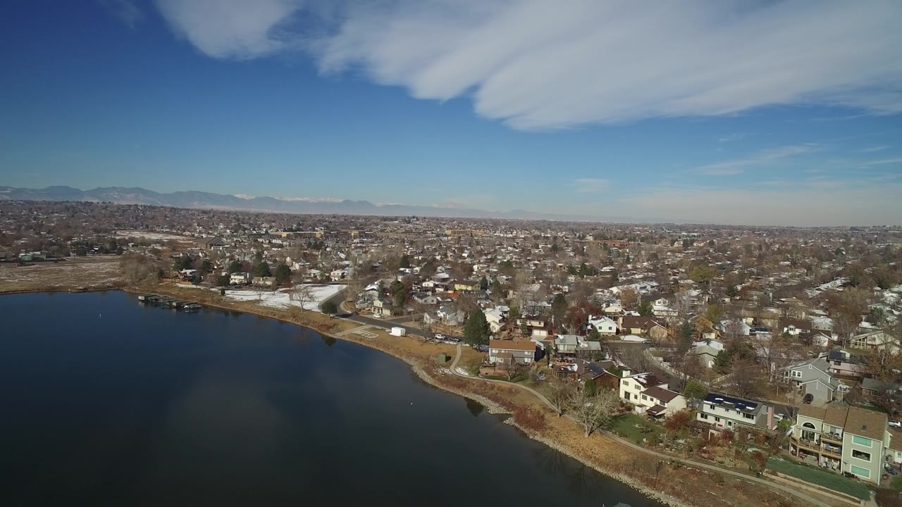 Aerial view in Hidden Lake, Colorado, showing more of the curved shoreline, clustered homes, leafless trees, patches of snow, distant mountains, blue sky, and no visible people.