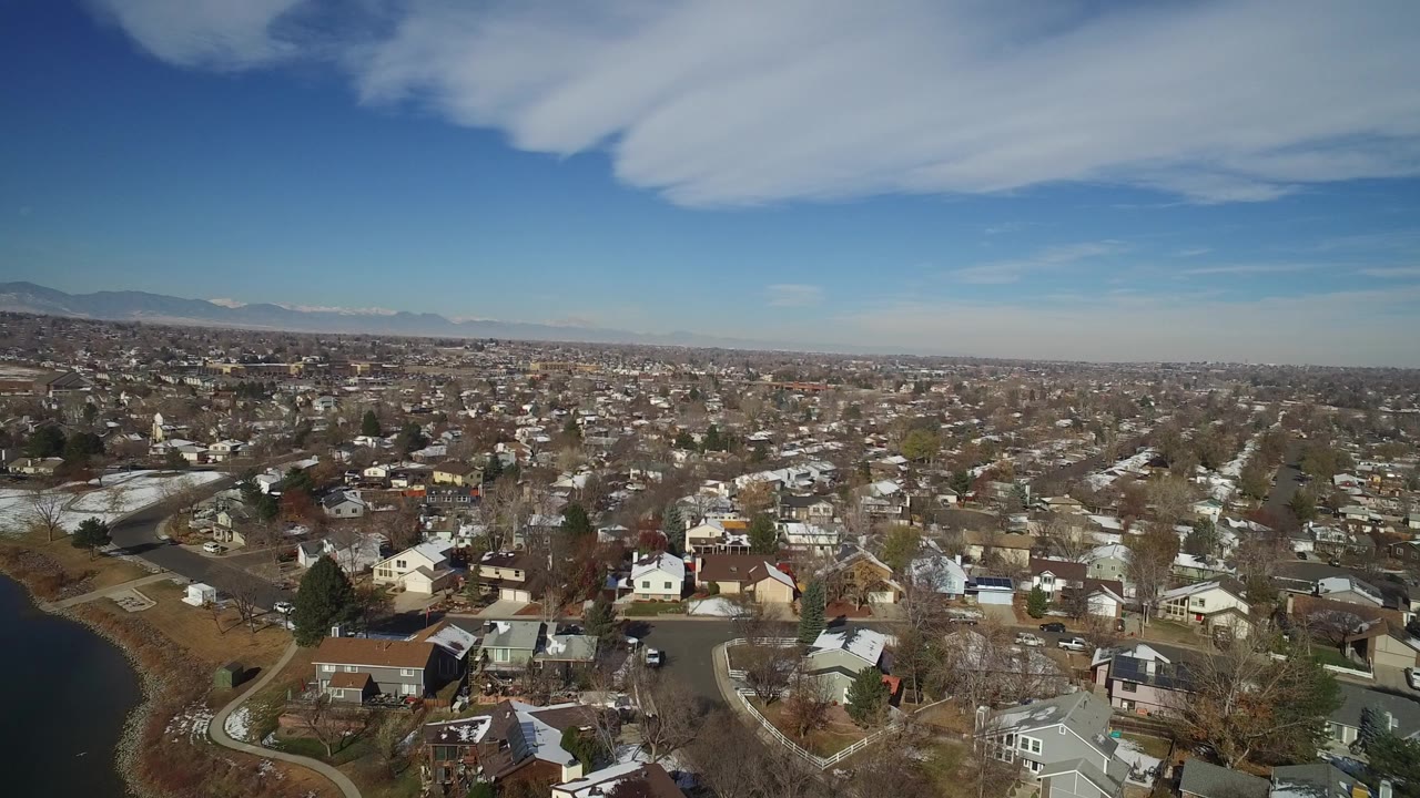 Aerial view over suburban houses, roads, trees, lake edge, blue sky, and distant mountains.