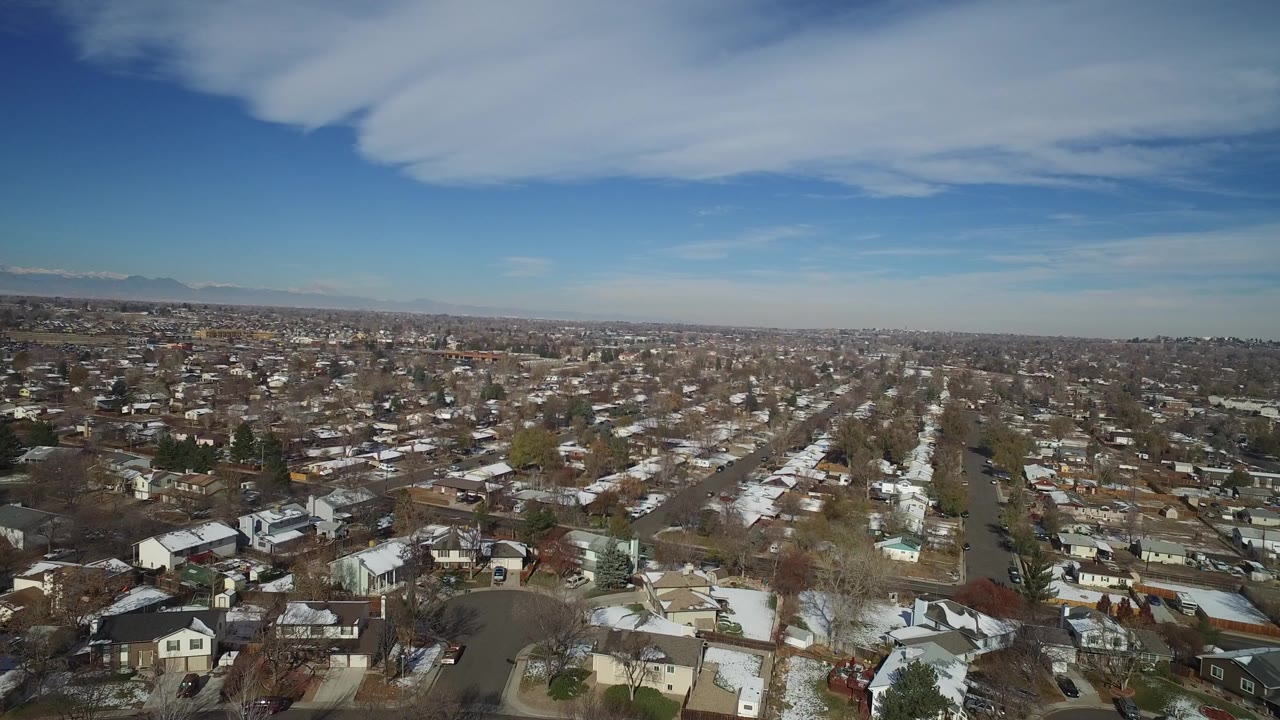 Aerial view of suburban homes, snowy lawns, and tree-lined streets under blue sky.