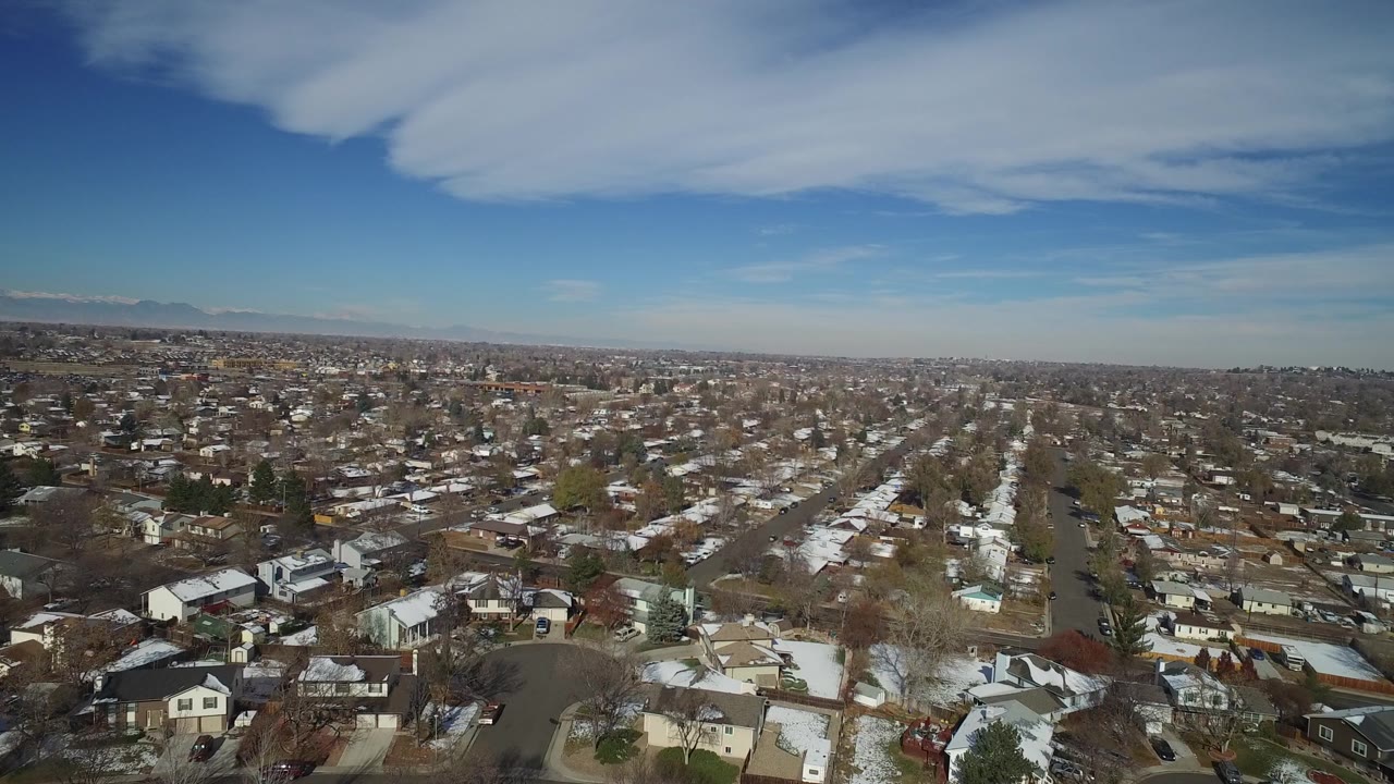 Wide aerial frame over Hidden Lake, Colorado with a long residential avenue, snow on roofs and yards, leafless trees, hazy distant hills, and no visible people.