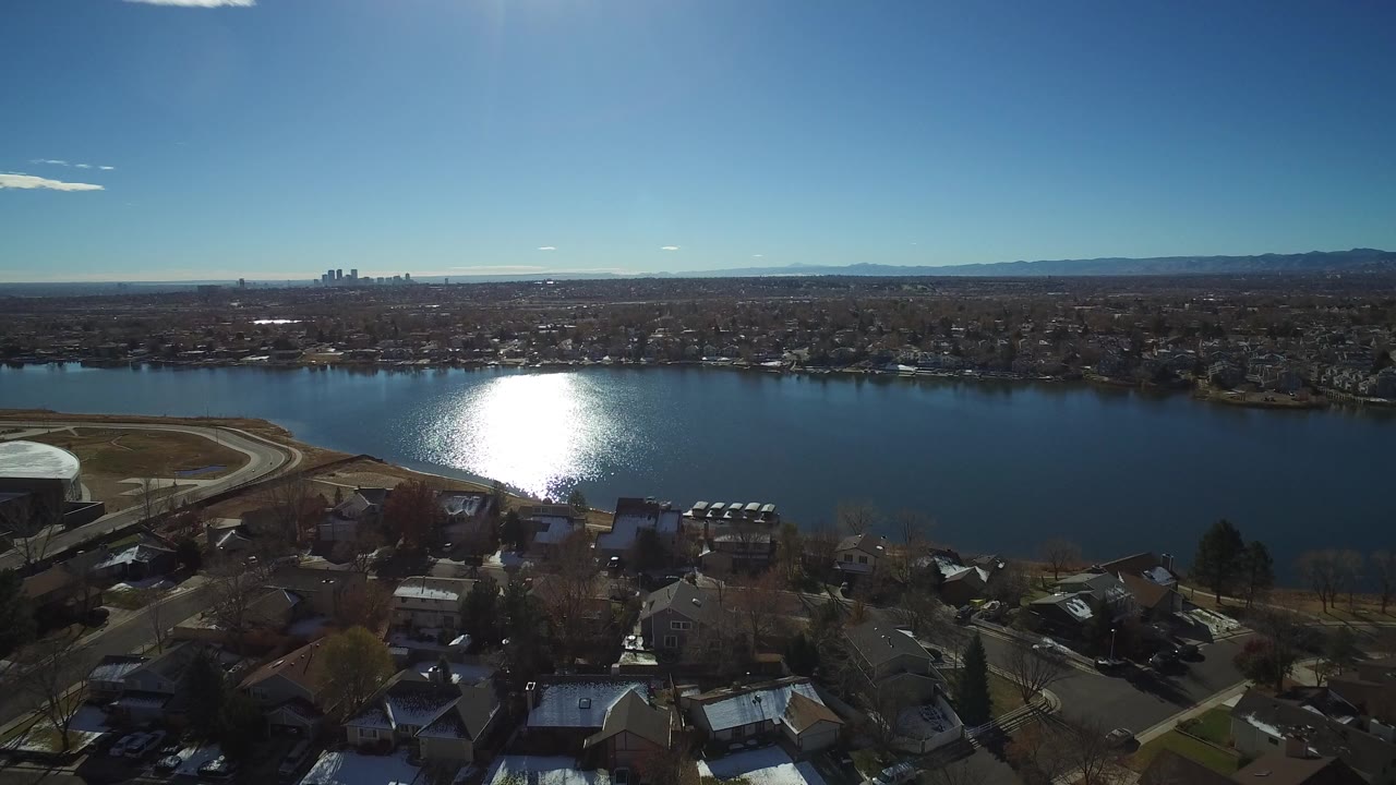 Aerial view of a lake with sun glare, houses, distant skyline, and mountains.