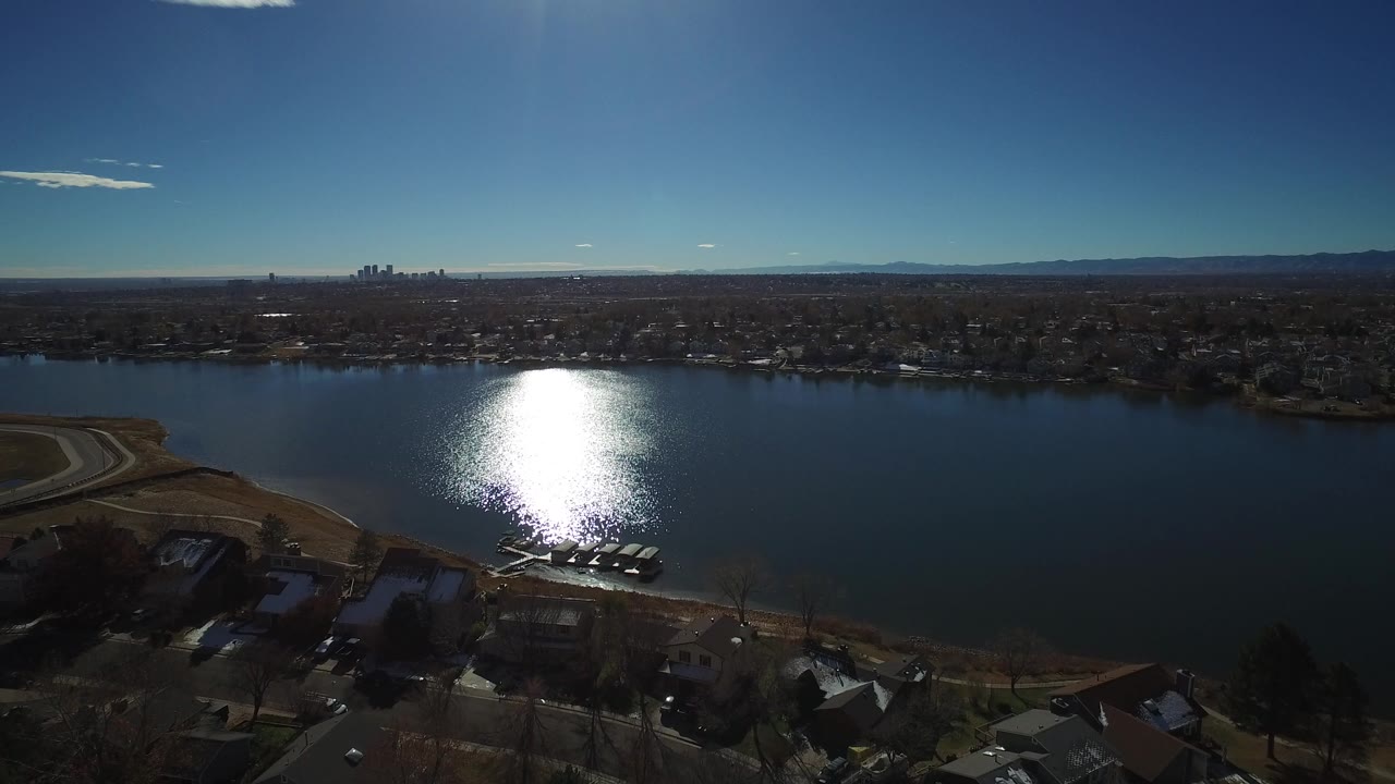 Aerial view over dark blue water toward suburban homes and a distant skyline.