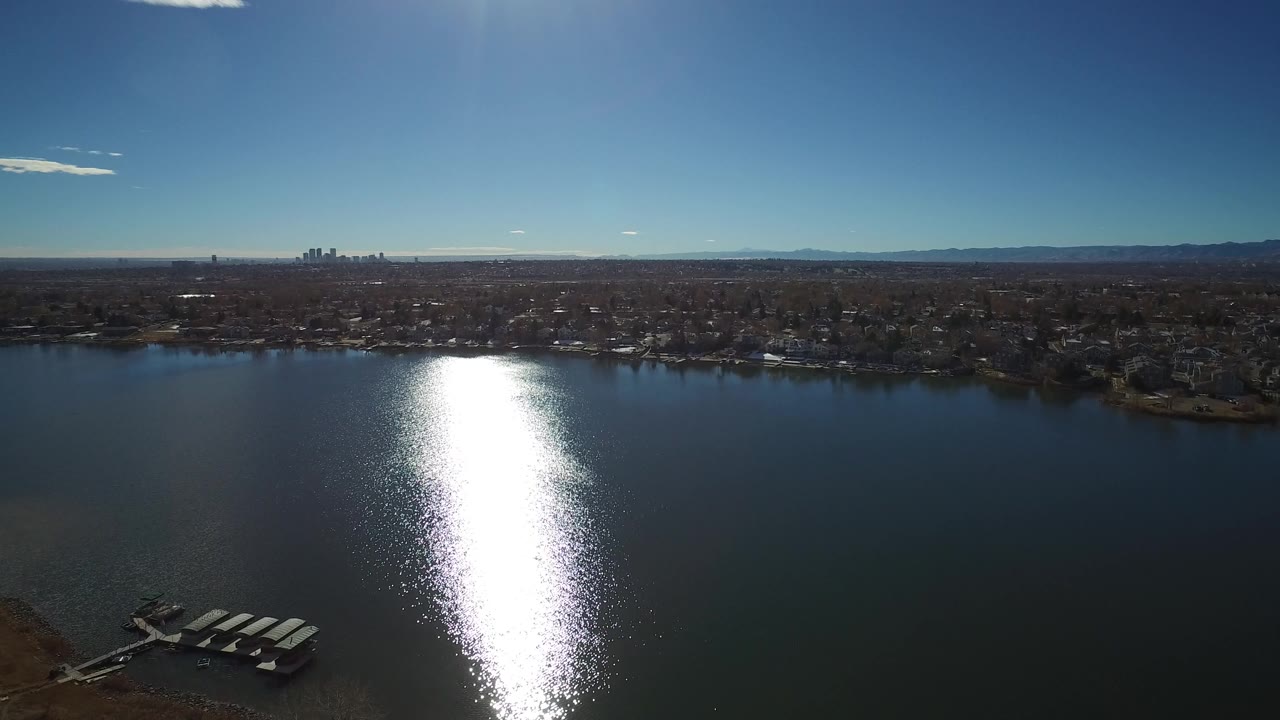 Wide aerial frame of a Westminster, Colorado waterfront, with the sun reflection centered on the lake, residential shoreline beyond, distant mountains, and no visible people.