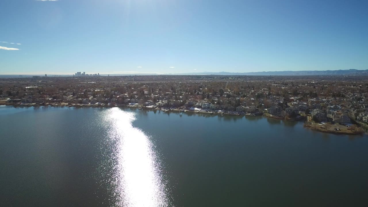 Wide aerial view over a lake with sun glare, shoreline houses, and distant mountains.