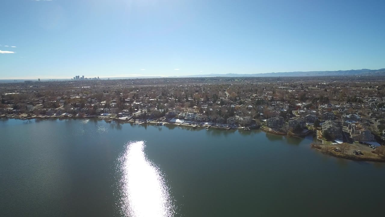 Aerial view over Westminster lake water with a tall patch of sun reflection, dense residential blocks along the shore, distant skyline at left, and no visible people.