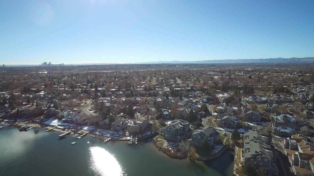 At 0:01 the Westminster, Colorado aerial keeps the lakefront homes low in frame, with dense residential blocks, bare trees, bright blue sky, and no visible people.