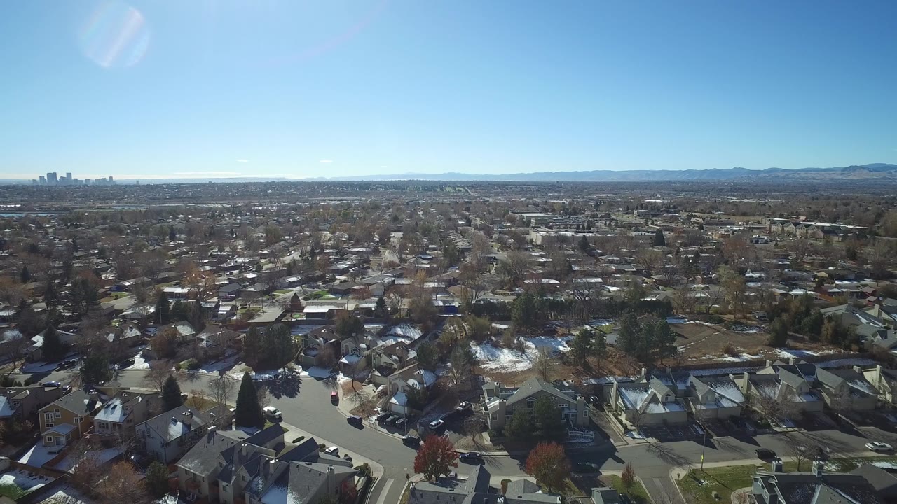 Aerial frame over Westminster, Colorado rooftops and curving streets, with patchy snow in yards, distant mountains across the horizon, and no visible people.
