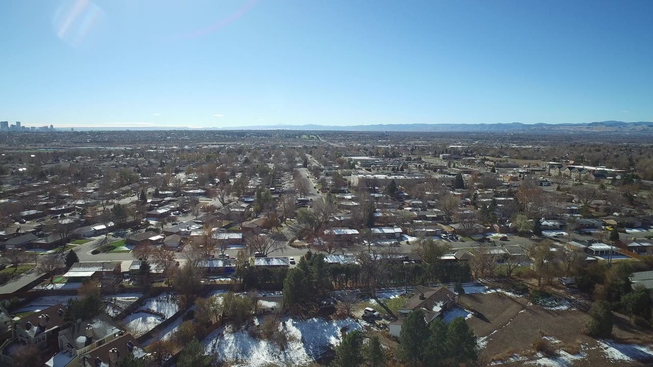Wide aerial frame over Westminster, Colorado suburbs with rooftops, bare trees, patches of snow, distant skyline, mountains on the horizon, and no visible people.