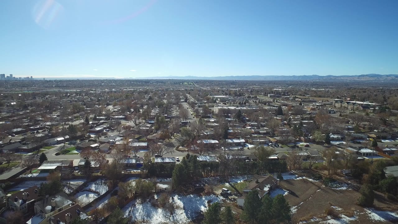 Aerial view across Westminster, Colorado residential blocks with a central street corridor, leafless trees, rooflines, far skyline, mountain backdrop, and no visible people.