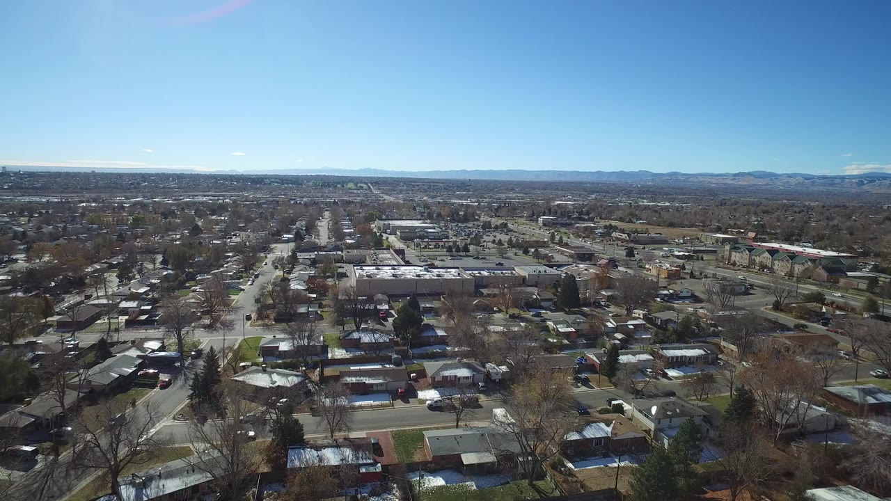Aerial frame over Westminster, Colorado showing residential rooftops, straight neighborhood roads, bare trees, distant mountains, and no visible people.