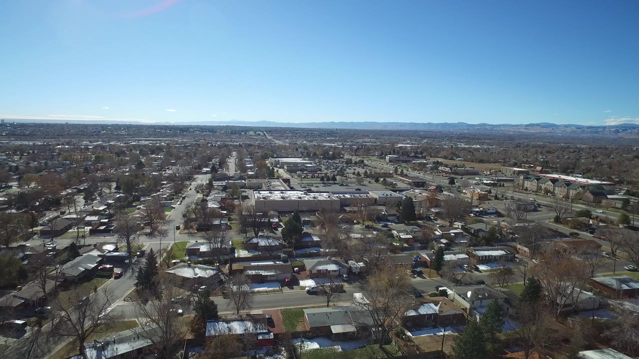 Aerial view in Westminster, Colorado with low-rise houses in the foreground, a long road corridor through the center, distant hills, and no visible people.