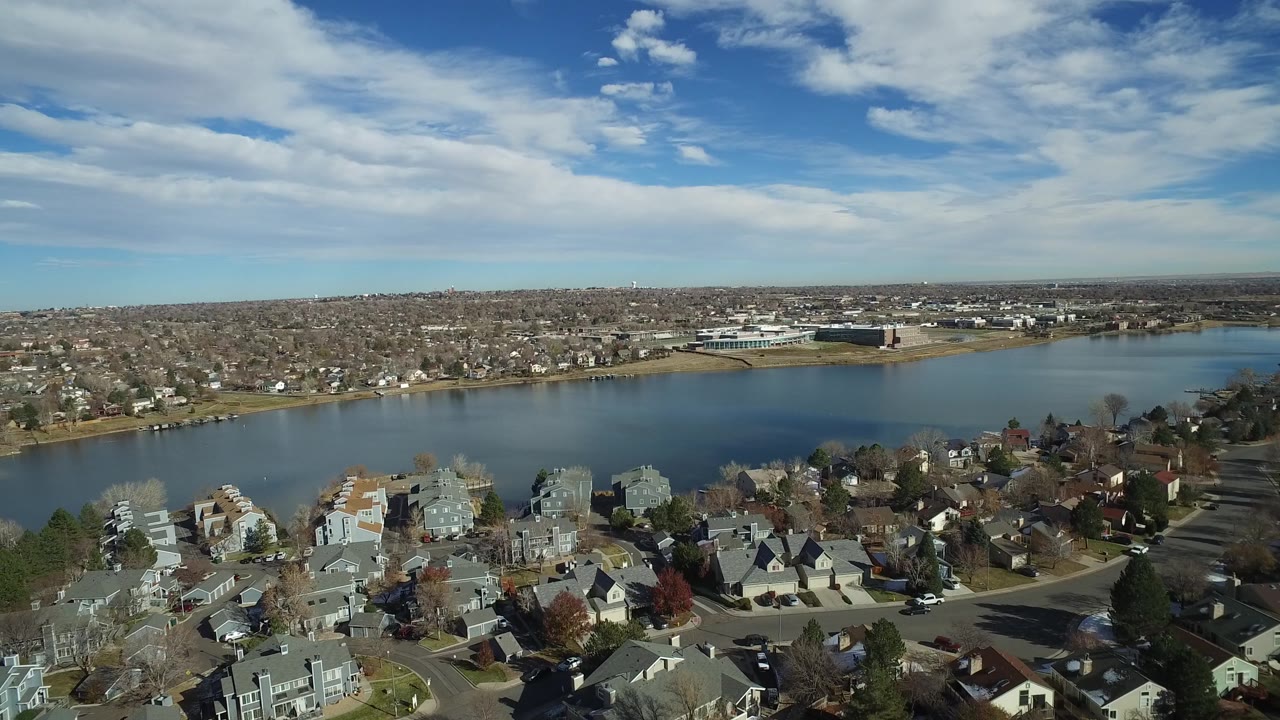 Westminster, Colorado aerial frame showing homes, bare autumn trees, a calm lake, low commercial-looking buildings in the distance, and no visible people.
