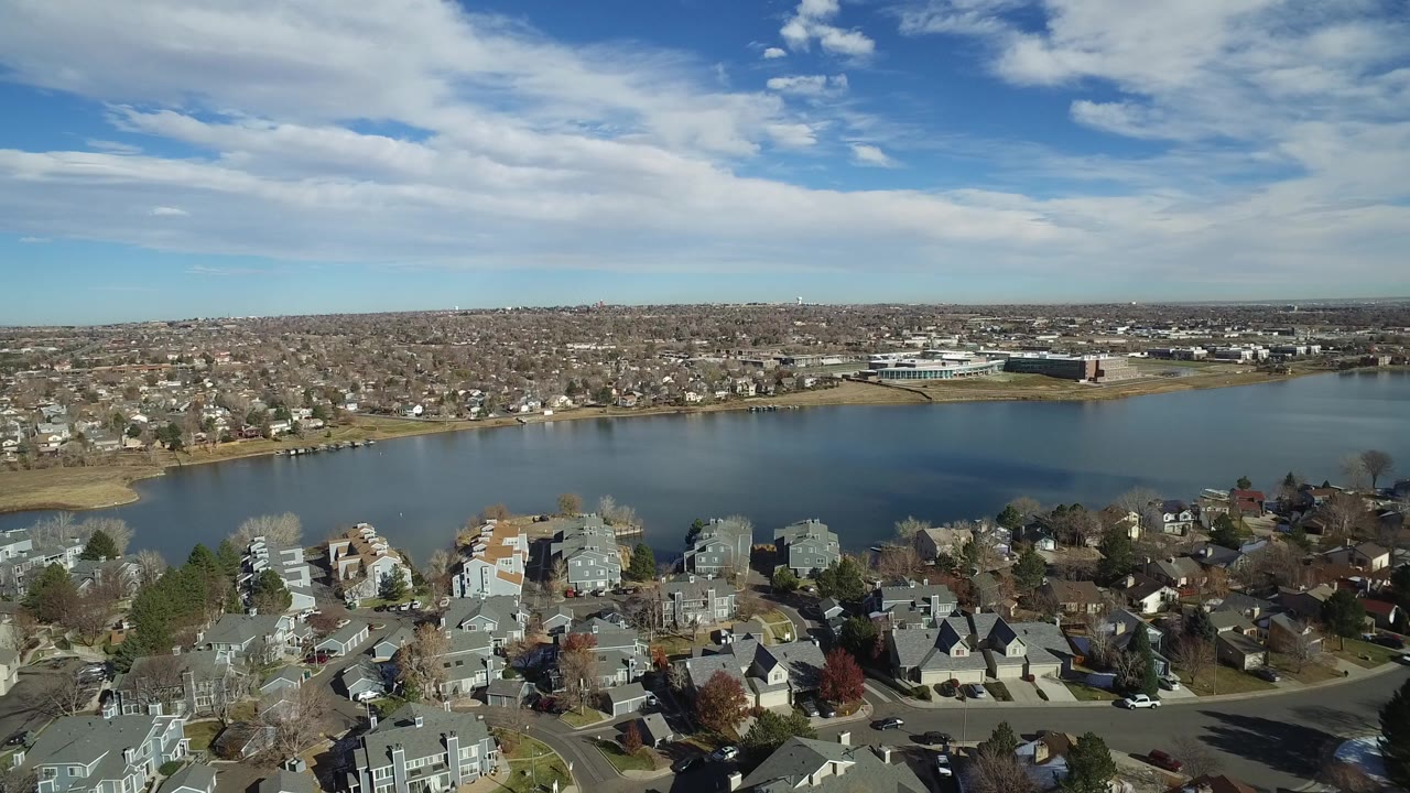 Opening aerial frame over Westminster, Colorado homes with a lake across the middle distance, low-rise city development on the horizon, and no visible people.