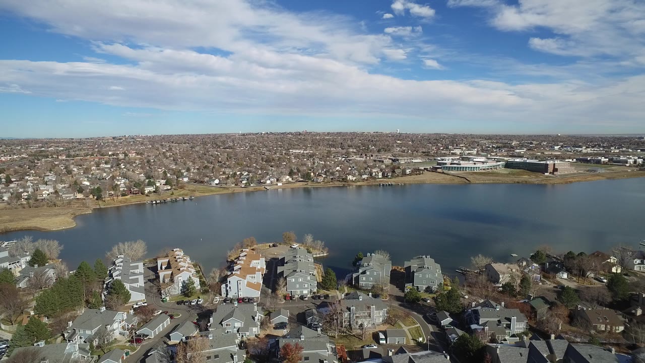 High aerial view in Westminster, Colorado with townhouses and parked cars in the foreground, a broad lake or reservoir across the middle, suburban homes on the far shore, and no visible people.