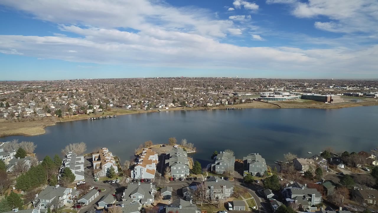 Drone-style aerial over lakeside homes with a suburban grid across the water.