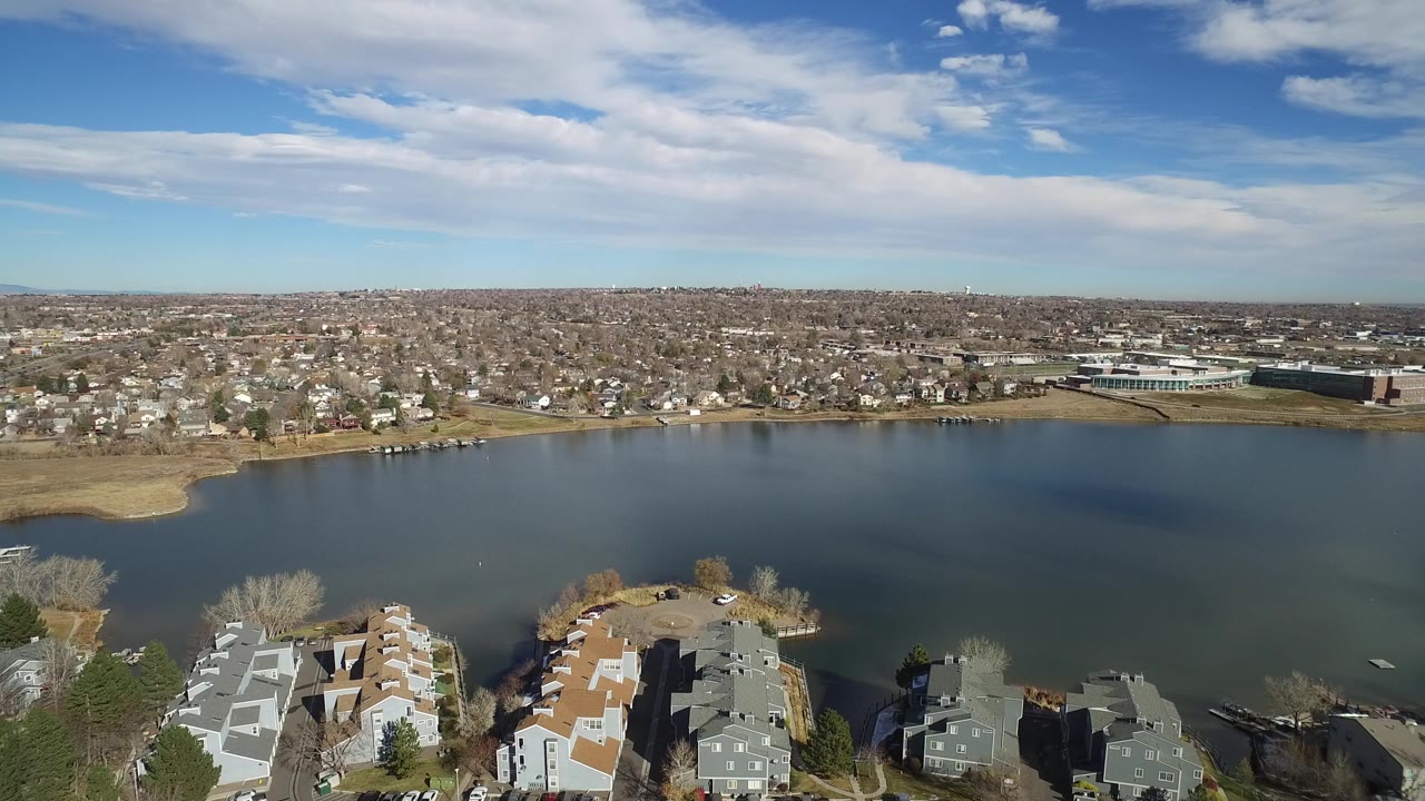 Aerial frame over Westminster, Colorado showing calm water centered in the image, clustered lakeside homes below, dense housing across the lake, and no visible people.