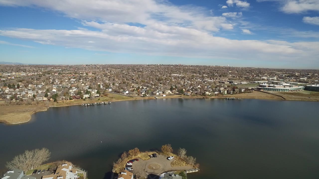 Aerial view in Westminster, Colorado looking across dark lake water to shoreline homes, a low building complex, distant hills, and no visible people.