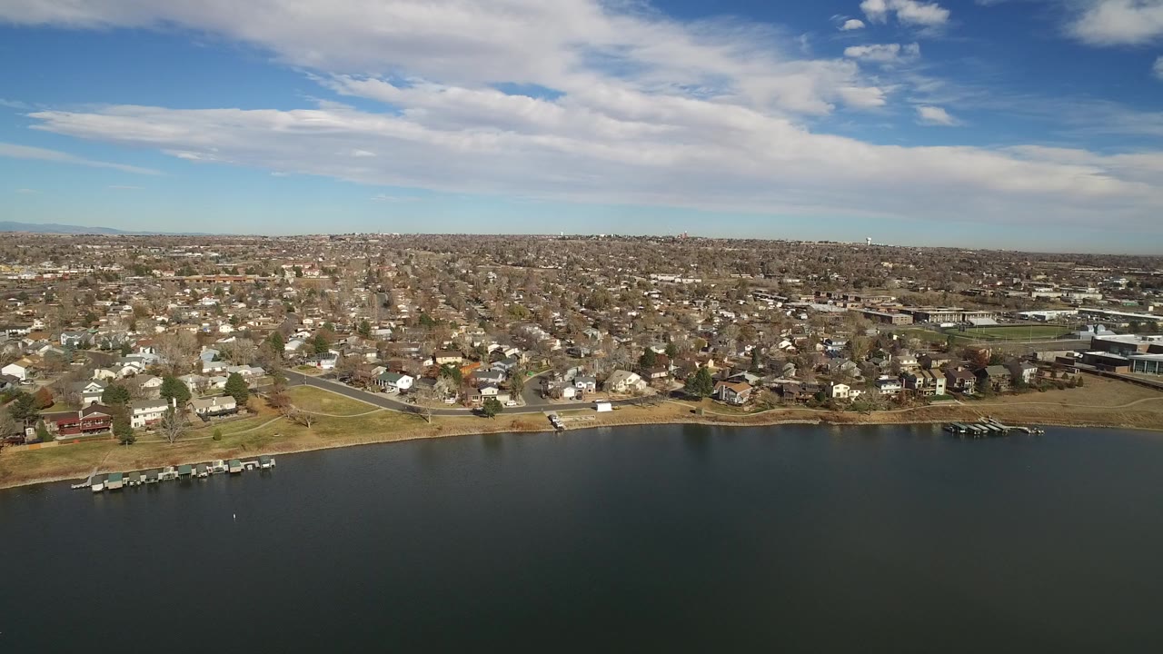 Aerial view of a lake shore and suburban neighborhood under a cloudy blue sky.