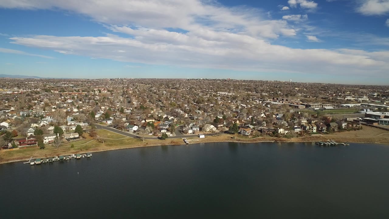 Aerial frame looking across the Westminster lakefront, with docks near the left shore, rows of houses beyond the water, and no visible people.
