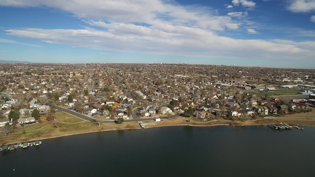 Wide aerial view of a lake, shoreline homes, and a suburban grid under clouds.
