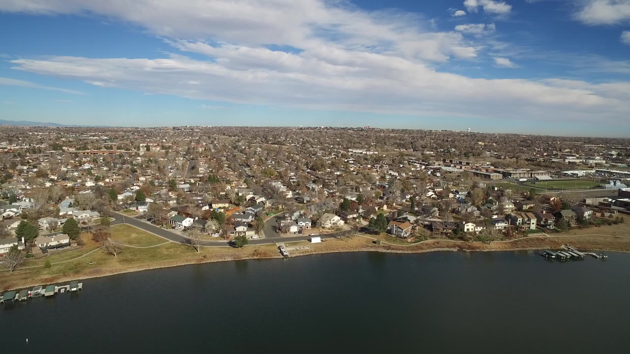 A Westminster, Colorado aerial still shows the lake edge across the lower frame, clustered homes and streets in the middle distance, a low horizon, and no visible people.