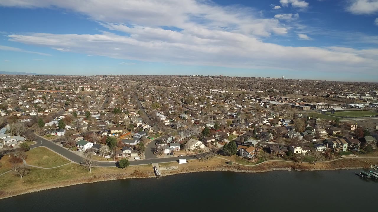 Opening aerial frame over Westminster, Colorado, with dark water in the foreground, shoreline grass, residential streets, and no visible people.