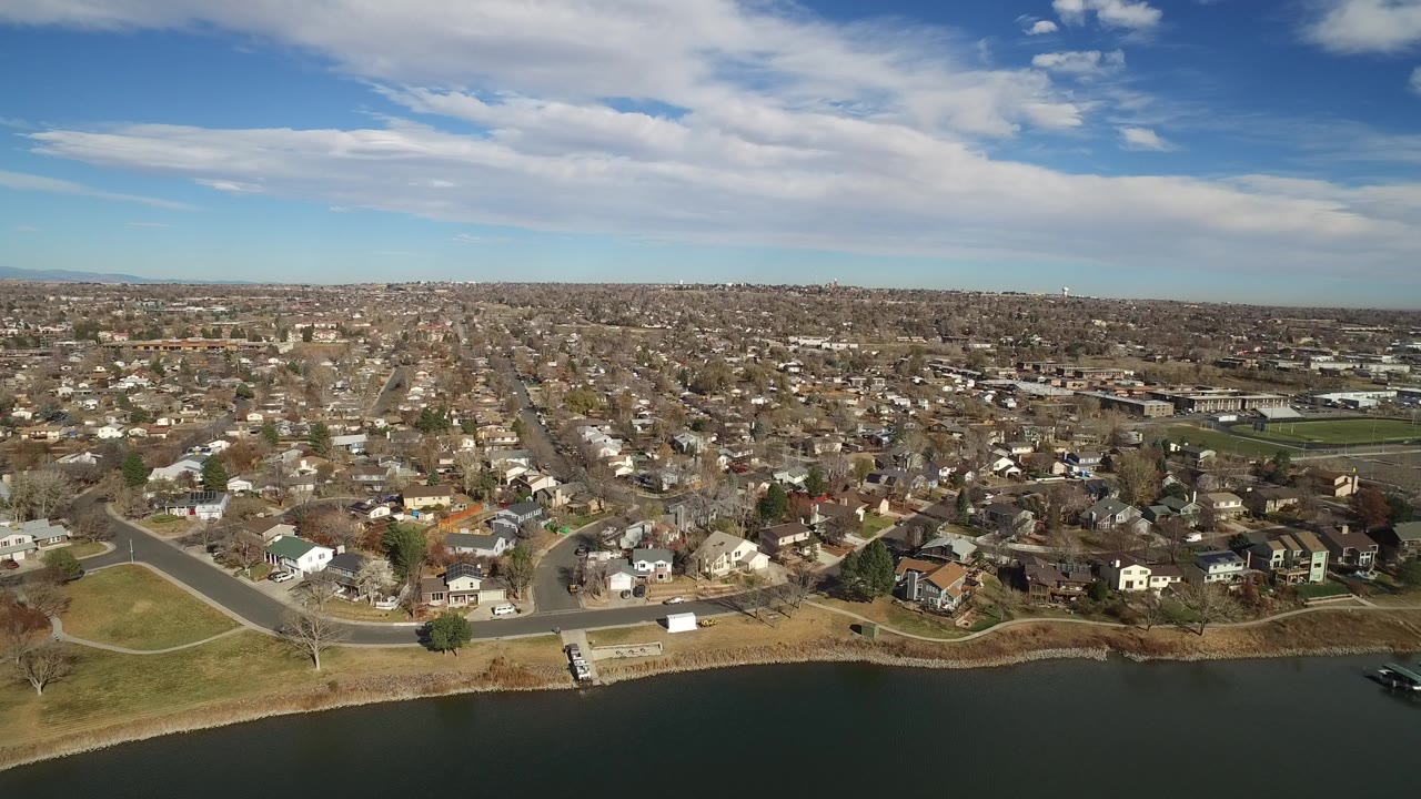 Wide drone view of houses, roads, and shoreline with a small dock area at the water.