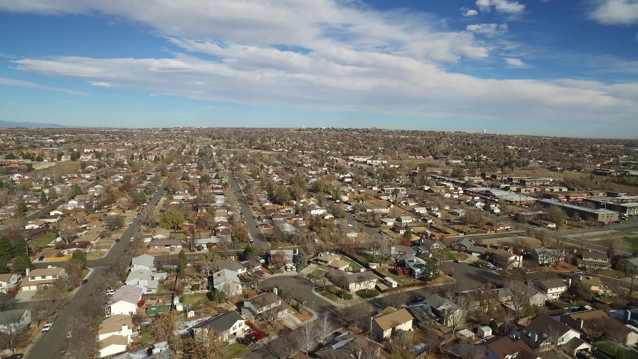 Wide aerial view over Westminster, Colorado homes and intersecting residential streets under a blue sky with long white clouds, with no visible people.