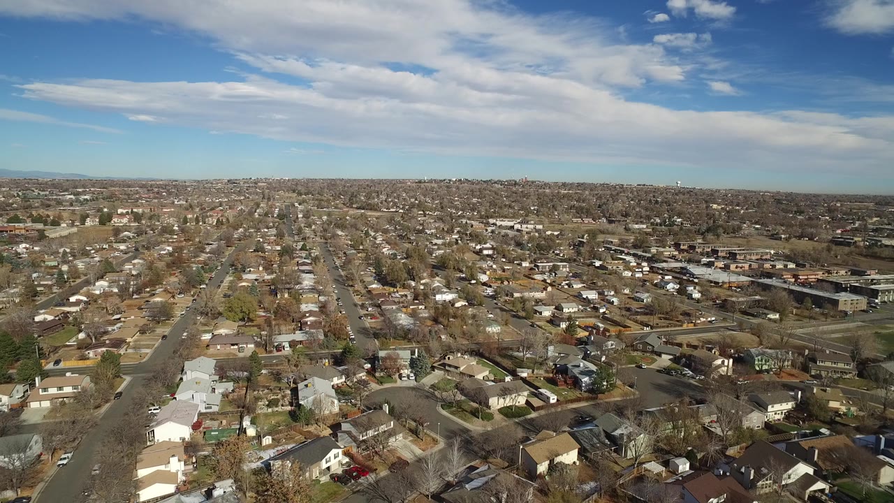 A high aerial frame shows Westminster, Colorado rooftops in the foreground, a straight neighborhood street running into the distance, scattered trees, and no visible people.