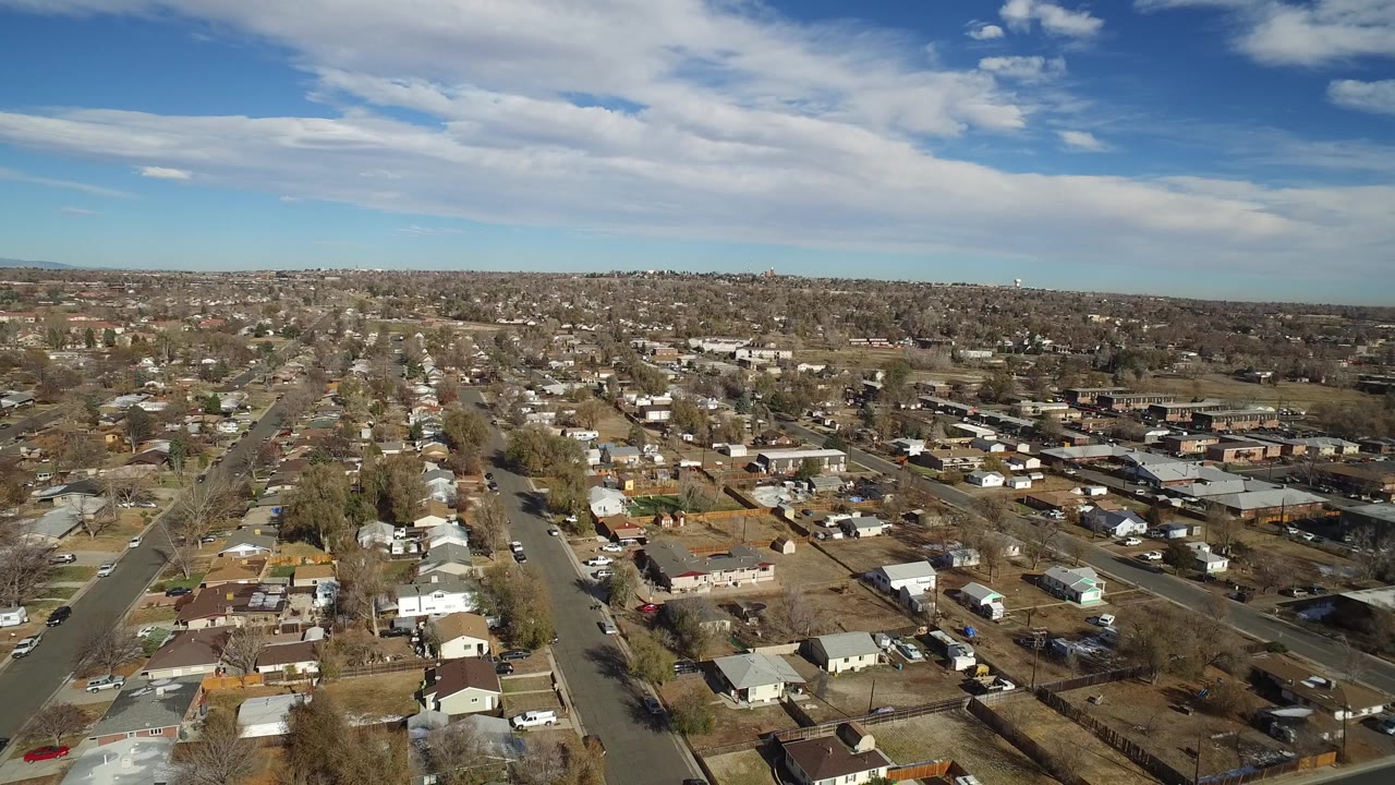Early aerial frame over Westminster, Colorado with a straight street running through houses, bare trees, backyard fences, and distant commercial roofs, with no visible people.