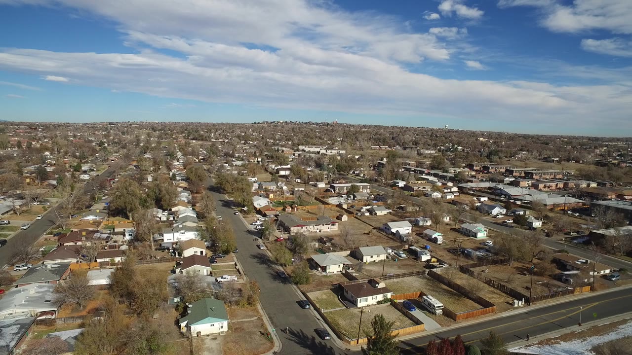 Aerial frame over Westminster, Colorado showing a residential street running through small homes, yards, leafless trees, parked vehicles, and no visible people.