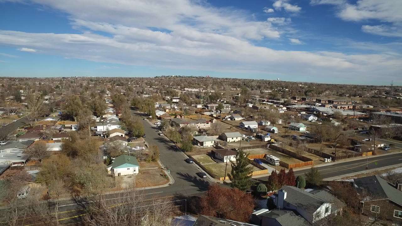 Early aerial frame over Westminster, Colorado homes, leafless trees, side streets, fenced yards, and a wide clouded sky with no visible people.