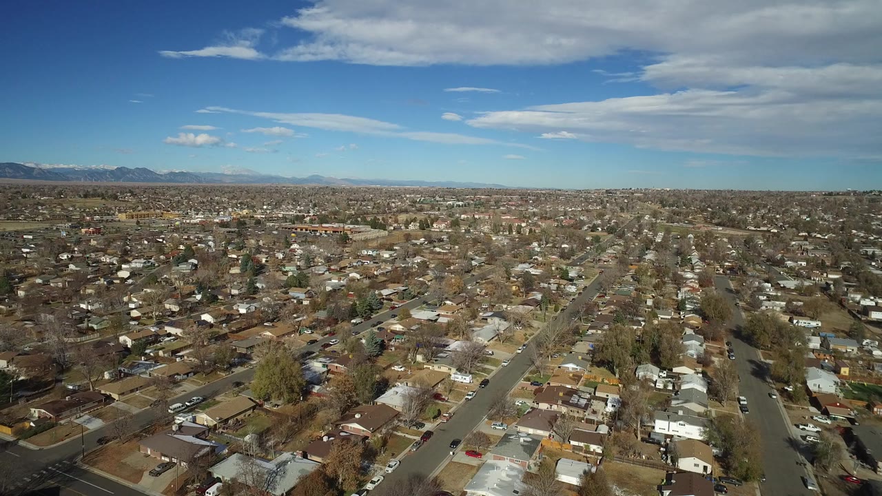 Westminster, Colorado neighborhood seen from above, with rows of homes, a long road through the frame, low winter trees, distant mountains, and no visible people.