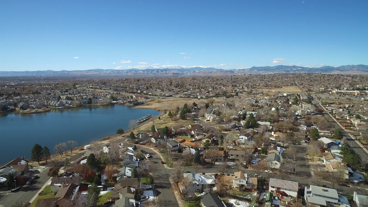 Aerial view over Westminster, Colorado homes and streets beside a blue lake, with distant mountains on the horizon and no visible people.
