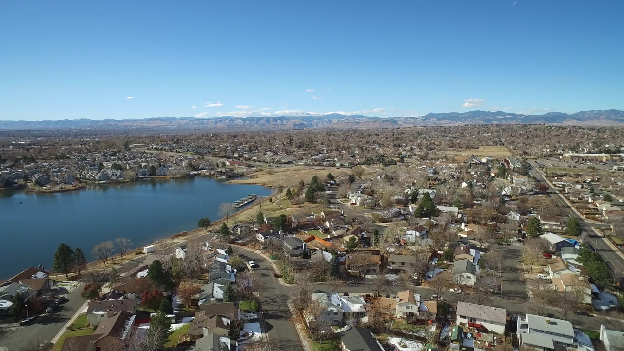 High aerial frame of Westminster residential blocks, lakeshore houses, and a wide blue lake, with the mountain range stretching across the background and no visible people.