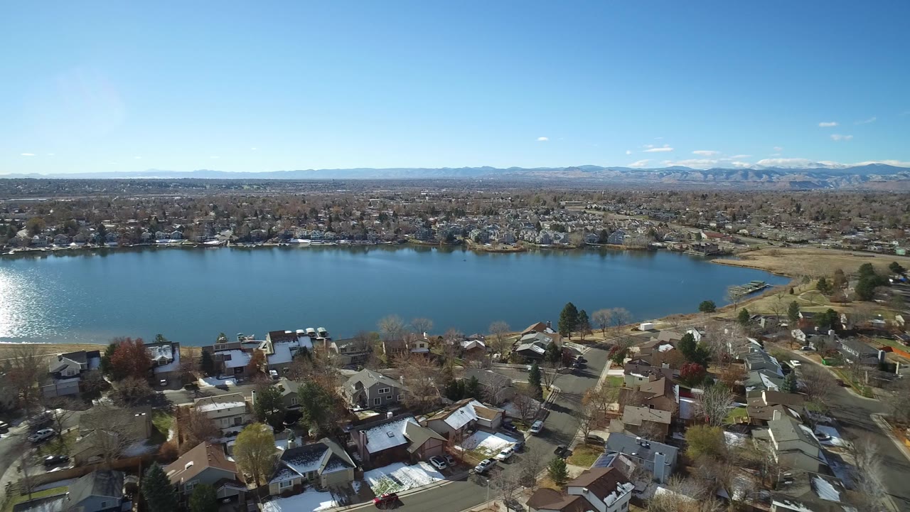 Aerial frame in Westminster, Colorado with bright sunlight reflecting on the lake, rows of homes and streets in the lower foreground, a far shoreline, and no visible people.