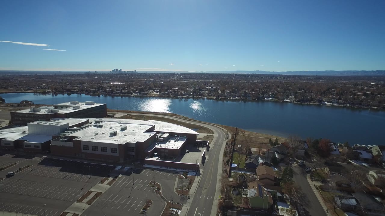 Wide aerial view in Westminster, Colorado showing a flat-roofed sports and school complex, parking lots, a river, distant skyline, and no visible people.